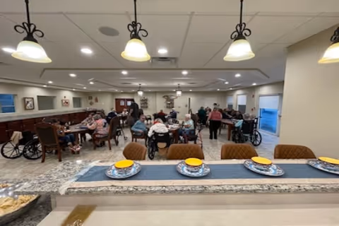 A dining room in a senior living facility with several elderly residents seated at tables, some in wheelchairs, engaging in conversation and activities. The room is well-lit with ceiling lights and hanging pendant lamps above a counter set with plates and bowls. The atmosphere appears communal and welcoming.