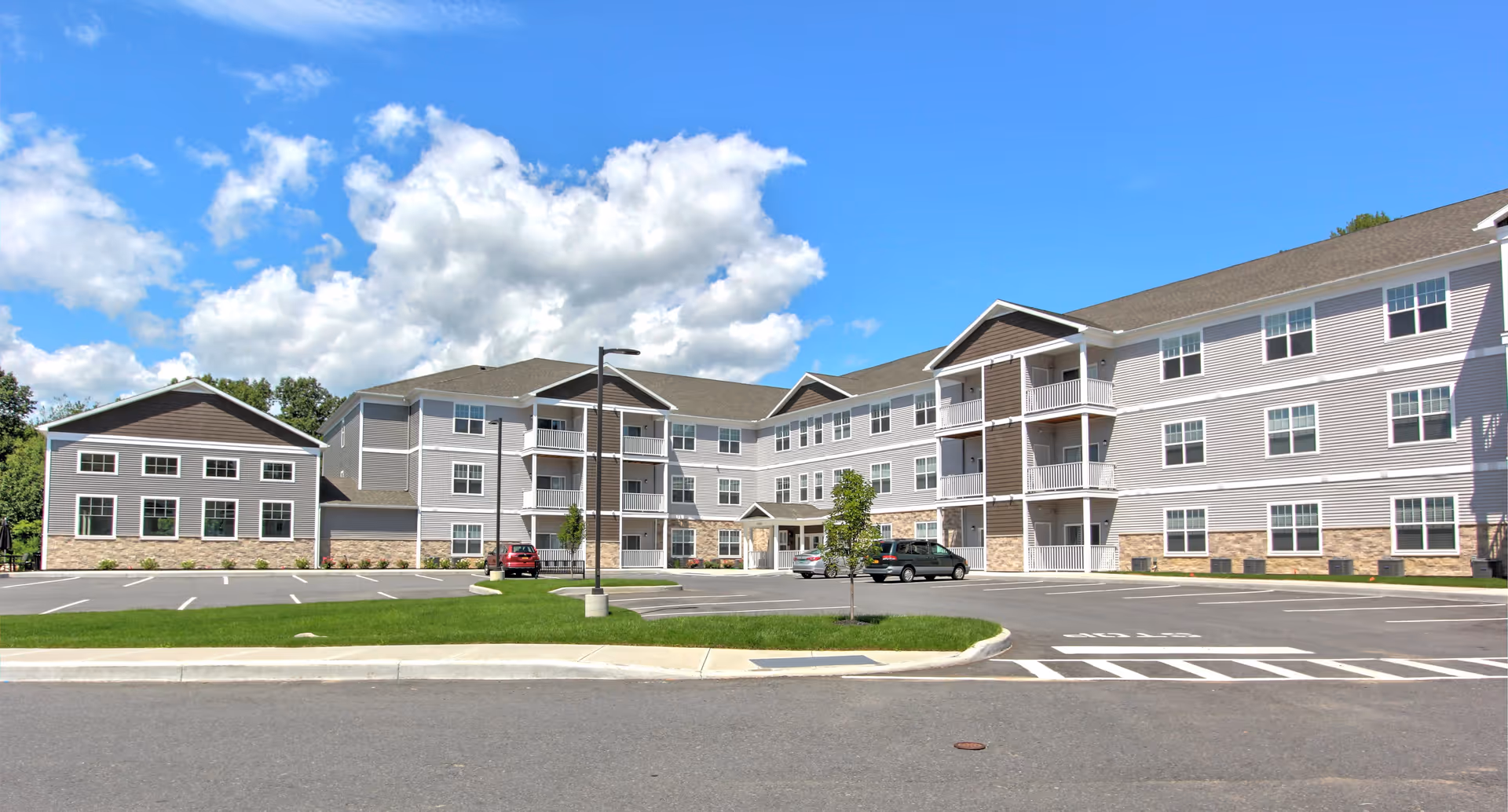 Exterior view of Carlton Hollow Senior Apartments, a three-story residential building with multiple windows and balconies, a parking lot with a few cars, green grass, and a bright blue sky with scattered clouds.