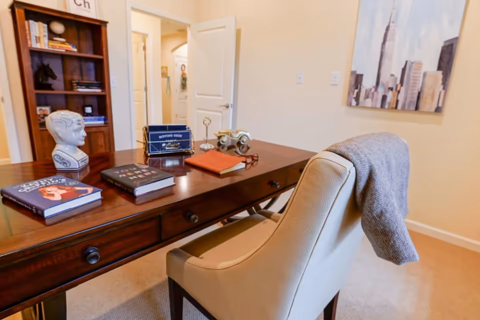 A cozy office or study room with a wooden desk holding books, a decorative head sculpture, a small model car, and a pair of glasses. A beige upholstered chair with a gray blanket draped over the back is positioned at the desk. In the background, there is a wooden bookshelf with books and decorative items, an open door leading to another room, and a large painting of a city skyline on the wall.