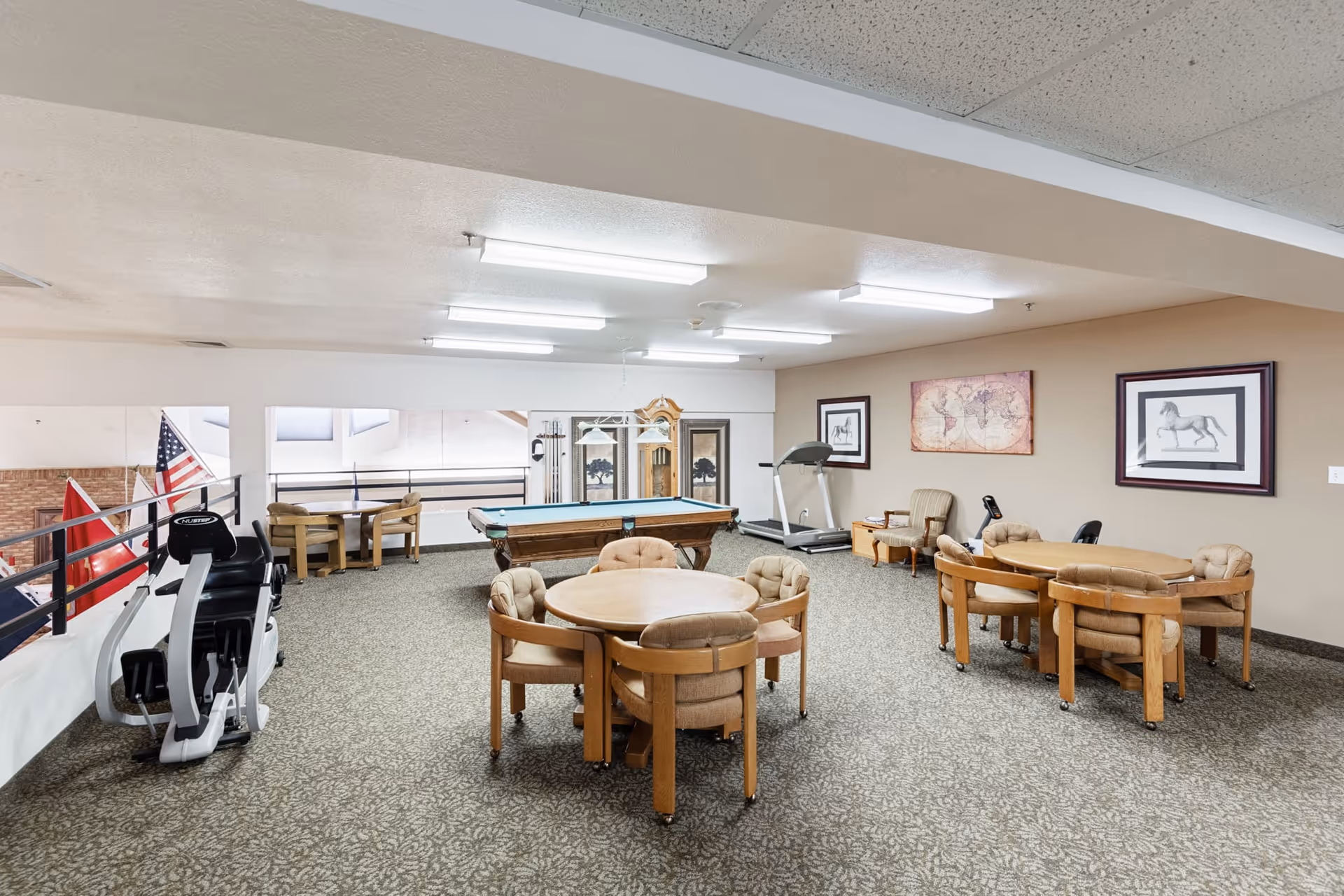A senior living community recreation room with round card tables and chairs, a pool table, exercise equipment, and framed artwork on the walls.