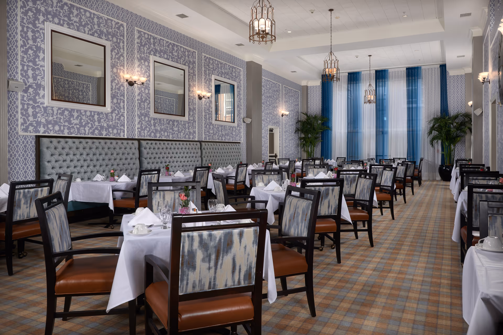 Elegant dining room with multiple tables covered in white tablecloths, each set with napkins, glassware, and small floral centerpieces. The room features patterned wallpaper, tufted bench seating along one wall, large windows with blue and white curtains, and hanging lantern-style light fixtures. The floor is carpeted with a plaid pattern.