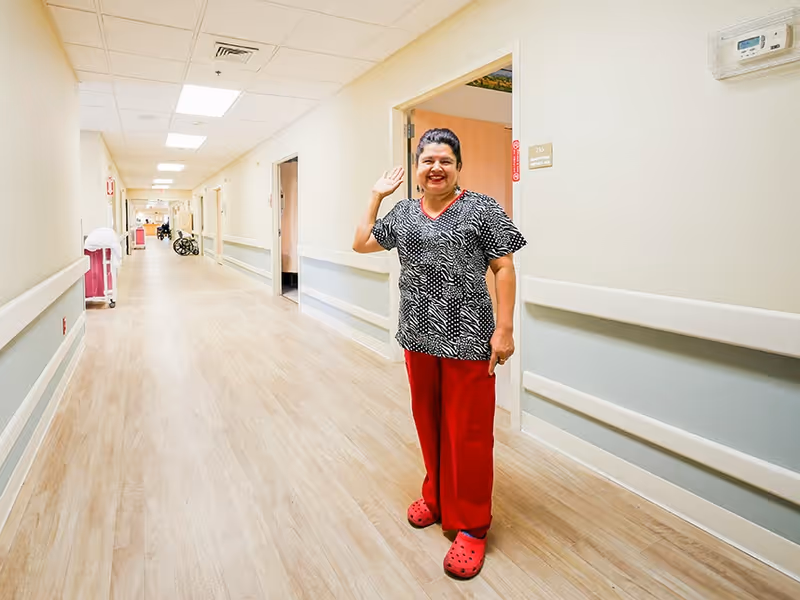 A smiling healthcare worker wearing a patterned black and white top, red pants, and red shoes is standing in a well-lit hallway of a healthcare facility, waving at the camera. The hallway has light-colored walls with handrails and several open doors along the corridor.