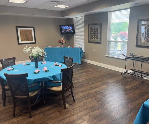 A dining room with a round table covered with a blue tablecloth and decorated with white flowers and pink napkins. There are six wooden chairs around the table. In the background, there is a rectangular table also covered with a blue tablecloth, a vase with flowers, a wall-mounted TV, two framed artworks on beige walls, and two windows letting in natural light. The floor is wooden.