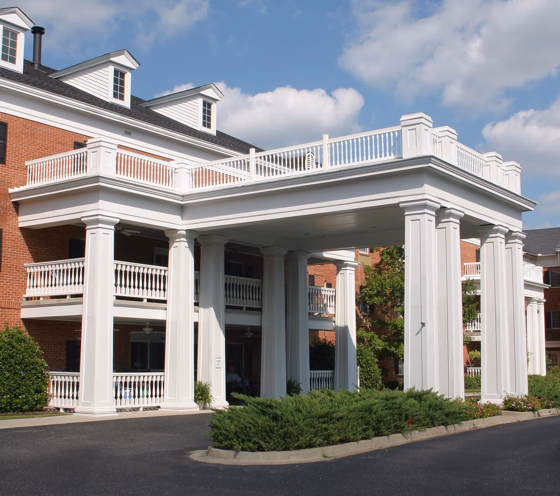 Entrance of a senior living facility with large white columns supporting a covered driveway. The building is made of red brick with white railings on balconies and dormer windows on the roof. There are shrubs and greenery around the entrance area under a partly cloudy sky.