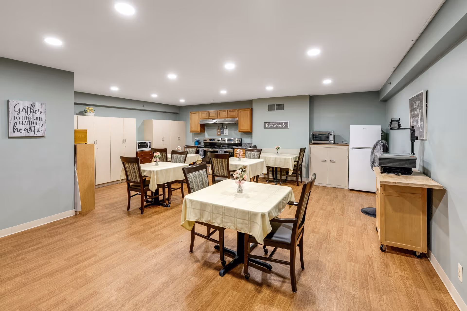 A dining area in a senior living facility with several tables covered with light yellow tablecloths and decorated with small flower vases. The room has wooden flooring, light green walls, and recessed ceiling lights. There is a kitchen area in the back with a stove, cabinets, a refrigerator, a microwave, and a toaster oven. Inspirational wall art is visible, including signs that say 'Gather together with grateful hearts' and 'Happiness is homemade.'