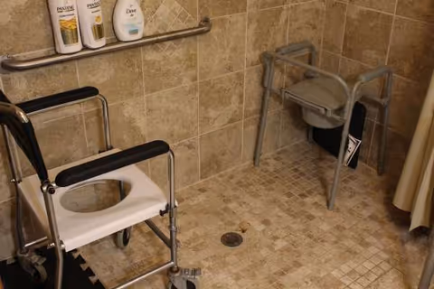 Accessible bathroom featuring a shower chair, grab bars, and a wheeled mobility chair.