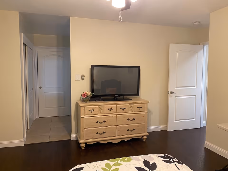 Bedroom showing a wooden dresser with a flat-screen TV, a portion of the bed in the foreground, and two open doors.