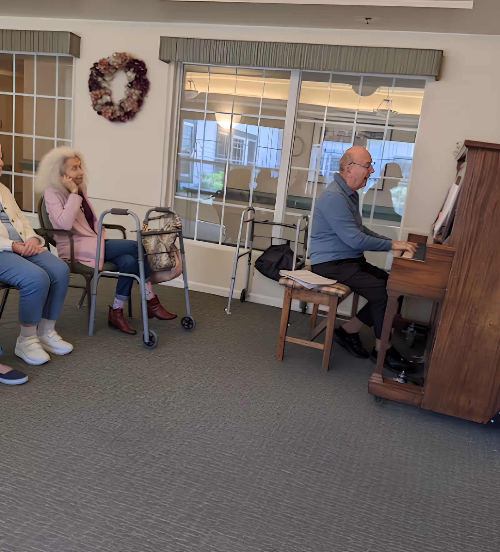 An elderly man playing a wooden piano in a room with large windows and a wreath on the wall. Two elderly women are seated nearby, one with a walker and a handbag, attentively listening to the music.