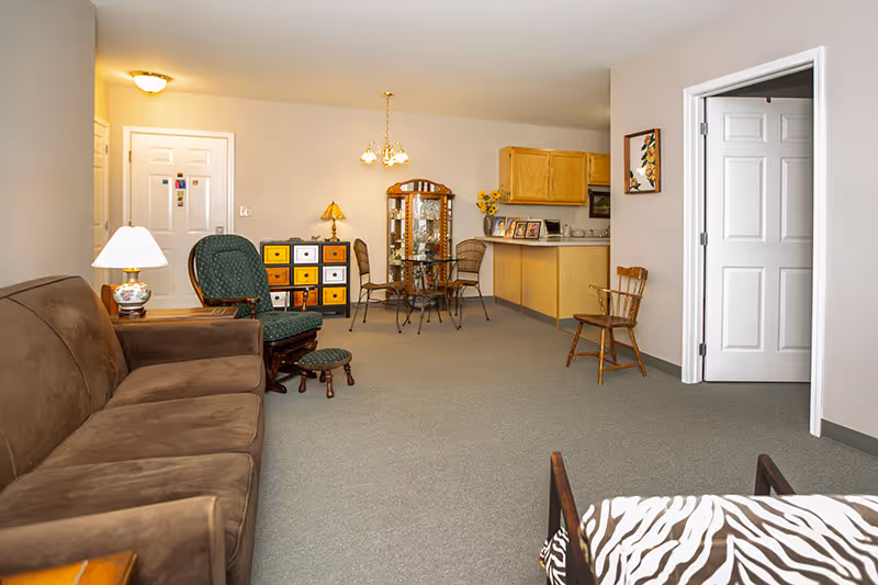 A spacious living area in an active adult community featuring a brown sofa, a green cushioned rocking chair with a matching footstool, a small wooden chair, and a glass-top dining table with four chairs. The room has beige walls, carpeted flooring, and a small kitchen area with wooden cabinets. There is a white door on the left side and an open doorway on the right.