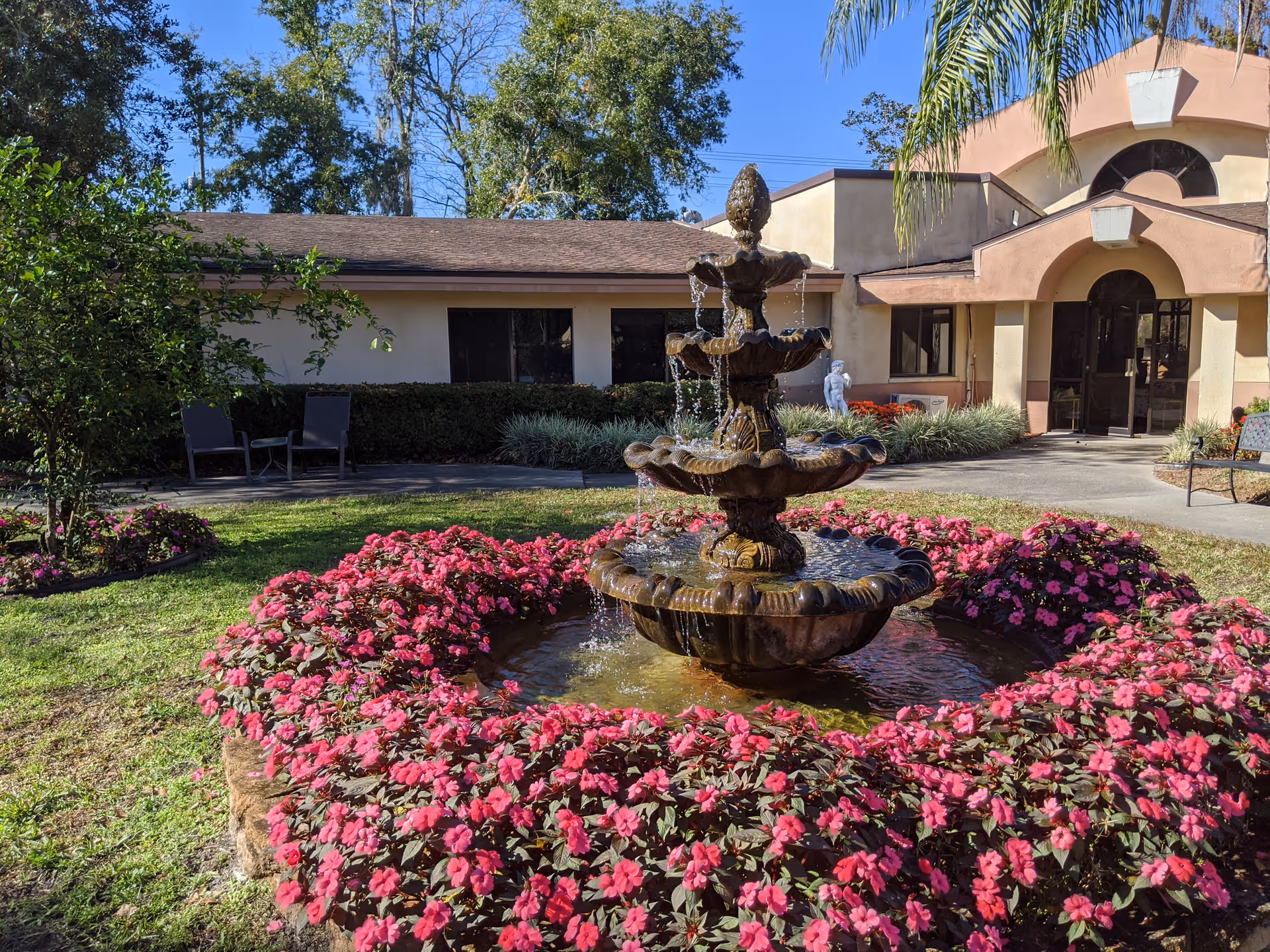 A three-tiered water fountain surrounded by a circular flower bed filled with pink flowers in a garden area. Behind the fountain is a building with beige walls, arched windows, and a brown roof. There are some chairs and a small statue near the building, with trees and greenery in the background under a clear blue sky.