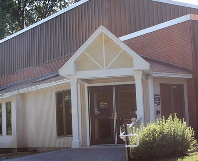 Front entrance of a community building with a peaked portico over double glass doors, brick and siding exterior, a handrail, and nearby shrubs.