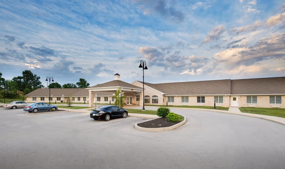 Exterior view of Avon Health and Rehabilitation Center showing a single-story building with a covered entrance, several parked cars, street lamps, and a partly cloudy sky.
