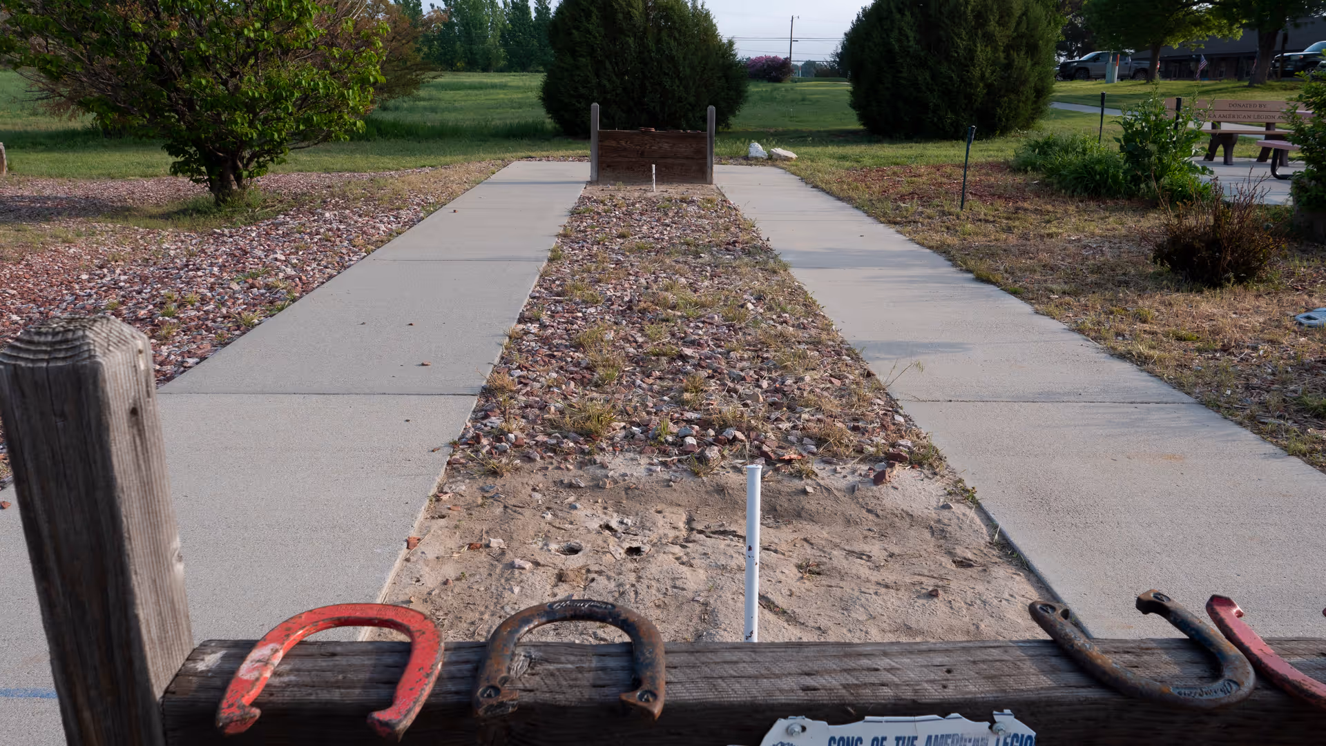 Outdoor horseshoe pitching area with a dirt and gravel lane, wooden backstop, and several horseshoes placed on a wooden beam in the foreground. Surrounding the area are grass, bushes, and trees under a clear sky.