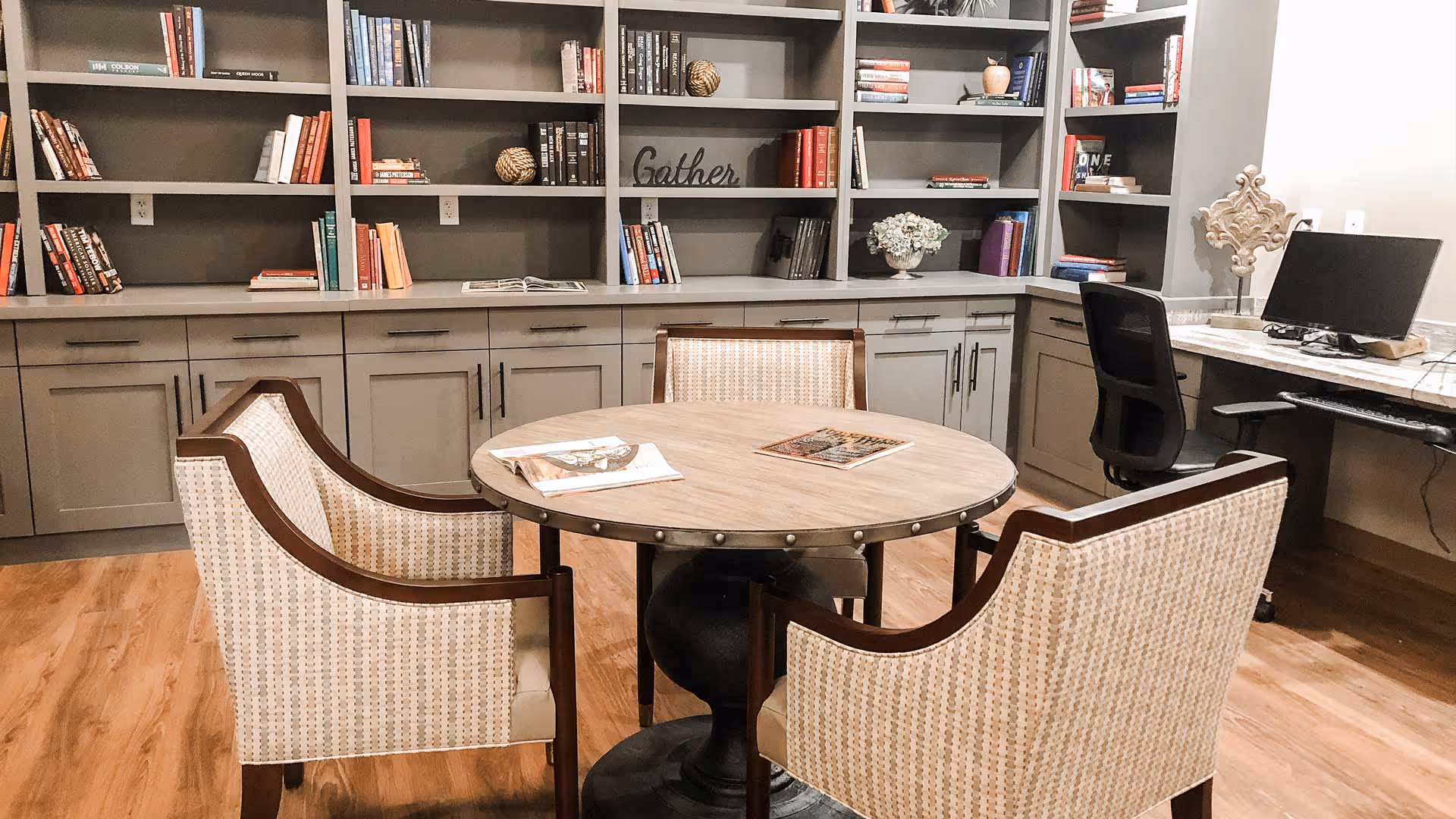 A cozy reading area featuring a round table with magazines, two patterned chairs, and a bookshelf filled with various books, creating an inviting atmosphere.