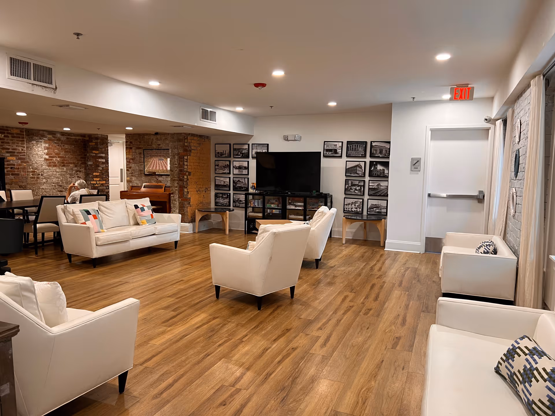 A spacious living room area in an assisted living facility with wooden flooring, white armchairs and sofas with colorful cushions, a flat-screen TV on a stand, and framed black-and-white photos on the wall. There is a dining area with tables and chairs in the background, exposed brick walls, and a person sitting at one of the tables. The room is well-lit with recessed ceiling lights and has an exit door on the right side.