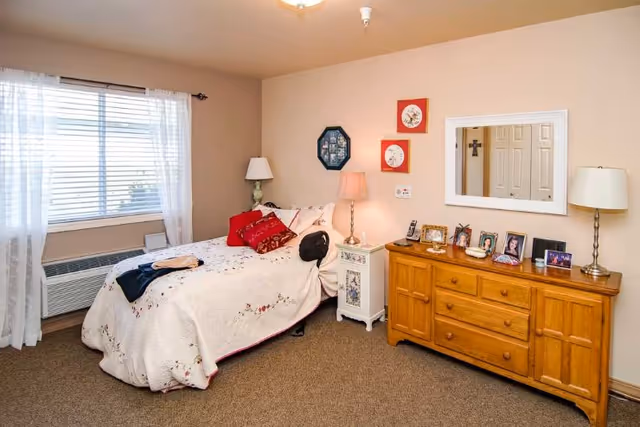 A cozy bedroom with a bed covered in a white floral quilt and red pillows, a wooden dresser with framed photos and decorative items, two lamps on either side of the bed and dresser, a window with sheer white curtains, and beige walls with framed artwork.