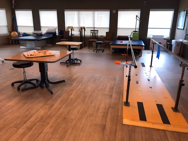 A rehabilitation therapy room with parallel bars for walking exercises, therapy tables, stools, and a table with a board game. The room has large windows with blinds and wood-patterned flooring.