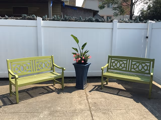 Two green metal benches facing each other with a tall blue planter containing green leaves and pink flowers placed between them, set on a concrete patio area enclosed by a white vinyl fence.