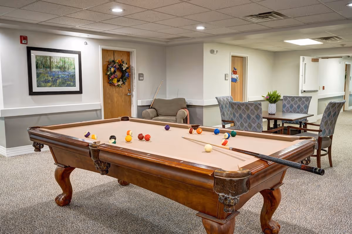 A recreational room in a senior living facility featuring a wooden pool table with billiard balls and cues on it. In the background, there is a small seating area with an armchair, a table with four patterned chairs, and a potted plant. The walls are decorated with a framed painting and a door adorned with a colorful wreath. The room has a carpeted floor and a drop ceiling with recessed lighting.