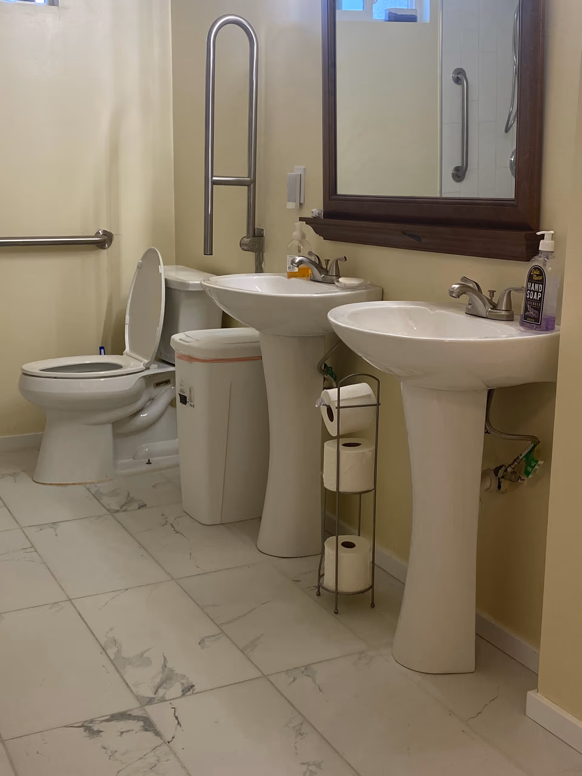 A bathroom with two white pedestal sinks, each with a faucet and soap dispenser. There is a large mirror above the sinks. To the left, there is a toilet with an open lid and a grab bar on the wall beside it. A white trash bin is placed between the toilet and the sinks. A metal stand holds three rolls of toilet paper. The floor is tiled with white and gray marble-patterned tiles, and the walls are painted light yellow.