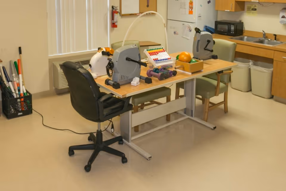 A room with a table holding two small pedal exercisers, an abacus, dumbbells, and a box of balls. There are three chairs around the table, one black office chair and two green cushioned chairs. In the background, there is a window with vertical blinds, a wall-mounted air conditioner, a fire extinguisher, a microwave on a countertop with a sink, and two trash bins underneath the counter. A basket with various sticks or rods is on the floor near the window.