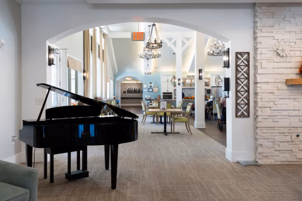 Interior view of a senior living facility showing a black grand piano in the foreground on the left, with a dining area in the background featuring tables and chairs. The space has high ceilings with chandeliers, large windows with curtains, and a white stone accent wall on the right.