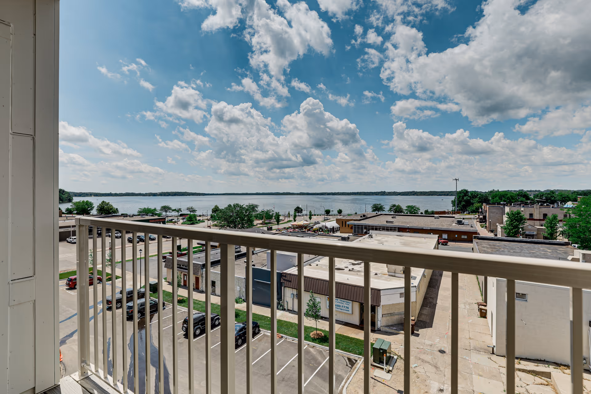 View from a balcony overlooking a parking lot, several buildings, trees, and a large body of water under a partly cloudy blue sky.