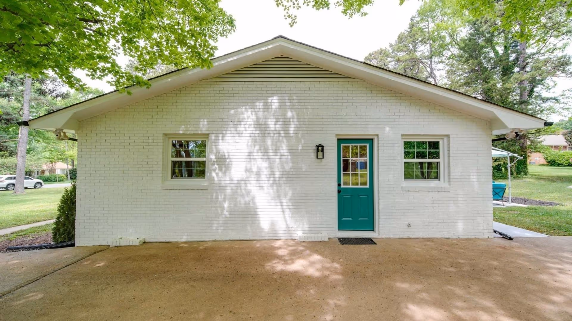 Front view of a small white brick building with a teal door and two windows, shaded by trees.