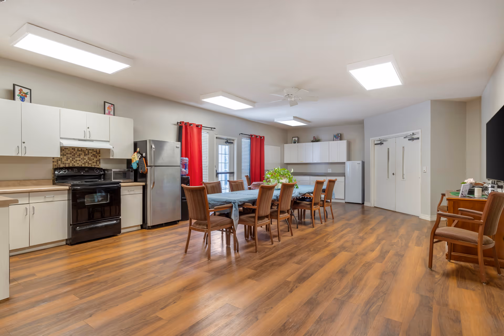 A spacious communal kitchen and dining area with wooden flooring, white cabinets, a black stove, a stainless steel refrigerator, and a water dispenser. There is a long dining table covered with a blue tablecloth surrounded by eight wooden chairs. Red curtains hang on the windows and door, and there are framed pictures on top of the cabinets. The room is well-lit with ceiling lights and a ceiling fan.