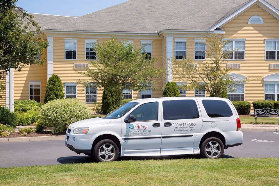 A white van with The Village at Buckland Court logo and contact information parked in front of a yellow two-story building with white trim, surrounded by green bushes and trees under a clear blue sky.