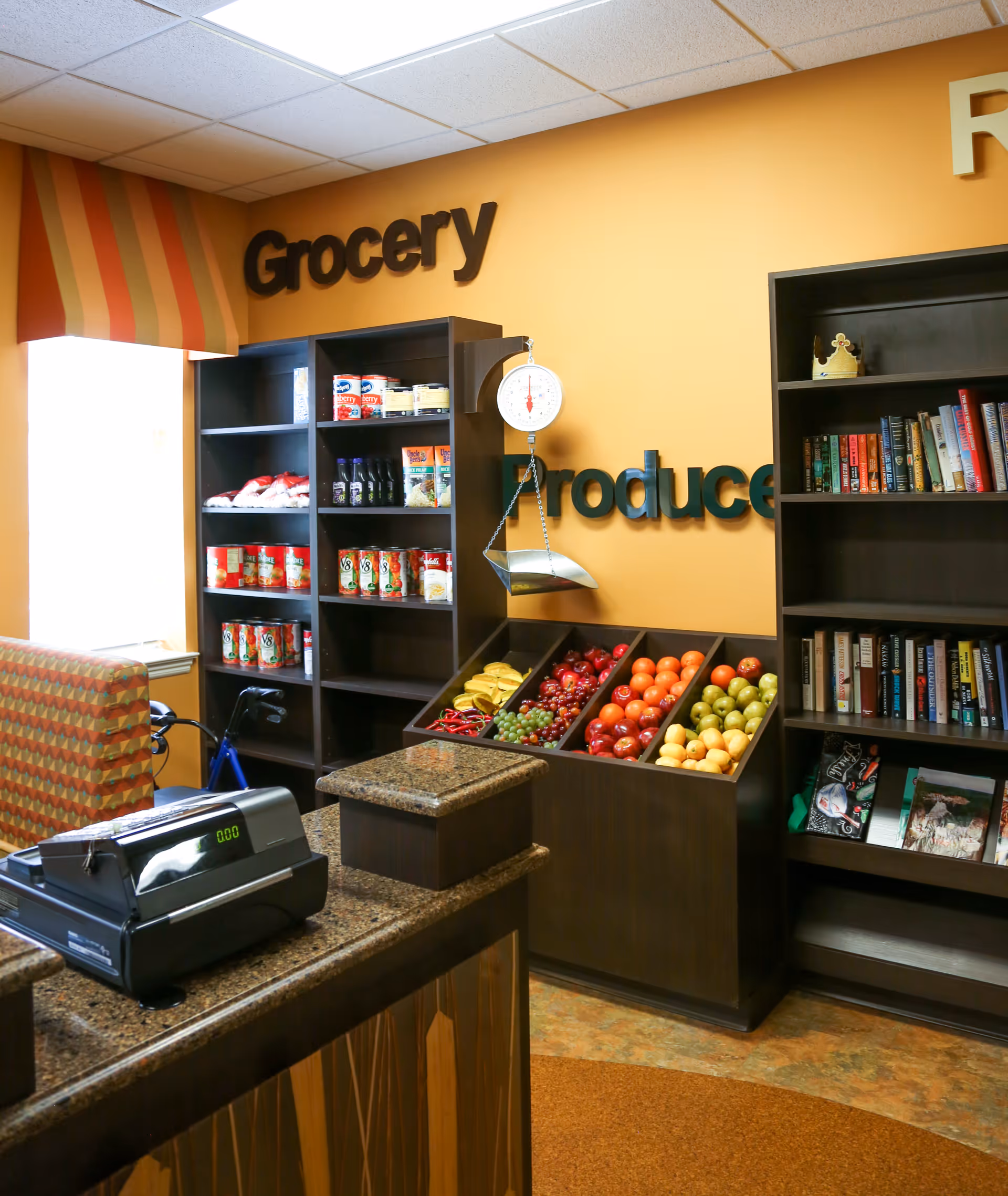 Interior view of a small grocery and produce section inside a facility. There are shelves stocked with canned goods and other grocery items, a produce display with bananas, grapes, apples, oranges, and pears, a hanging scale, a cash register on a counter, and a bookshelf with books and decorative items. The walls are painted orange with partial words 'Grocery' and 'Produce' visible.