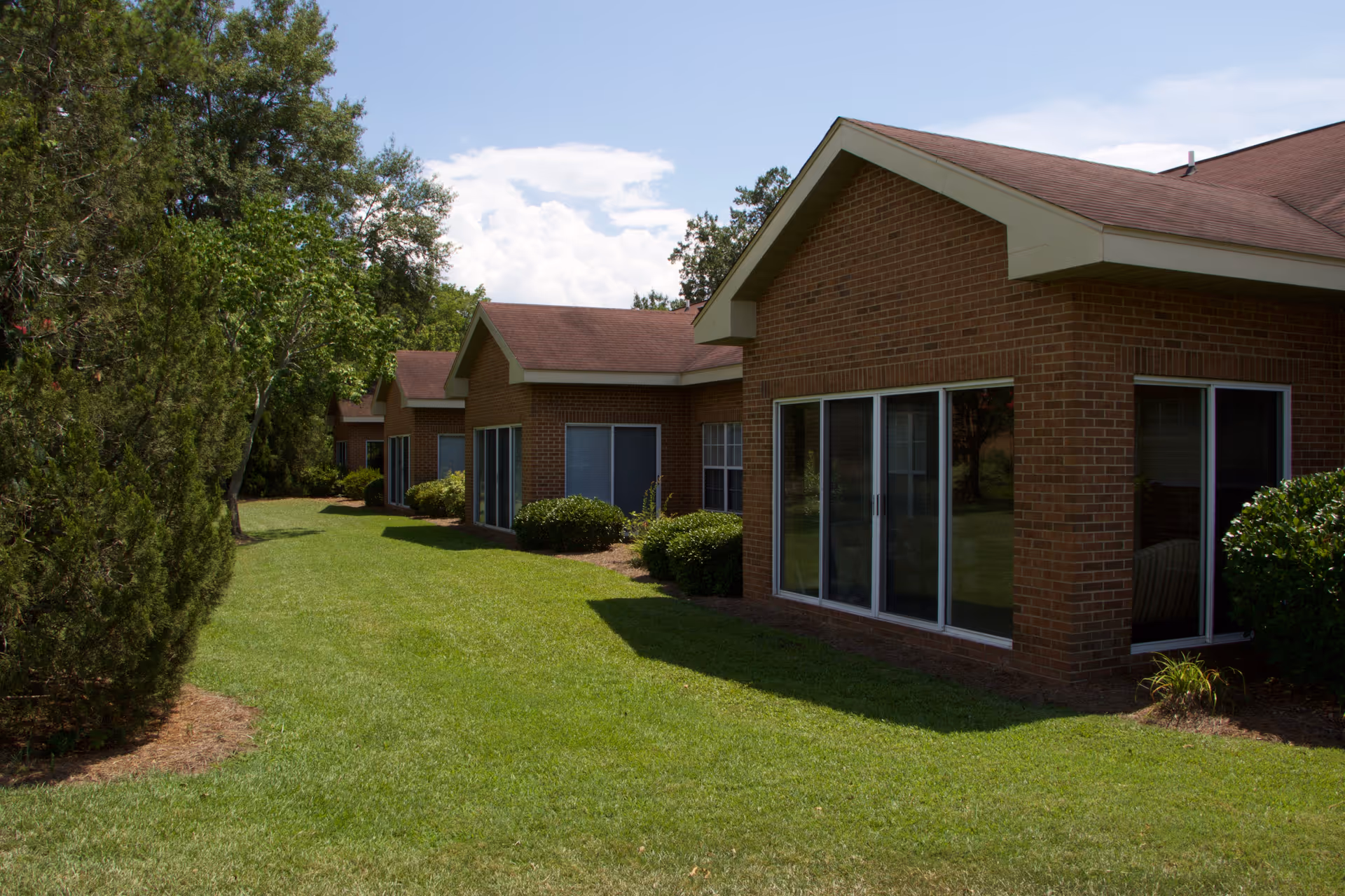 Exterior view of a single-story brick building with large windows and a well-maintained grassy lawn surrounded by trees and shrubs under a partly cloudy sky.