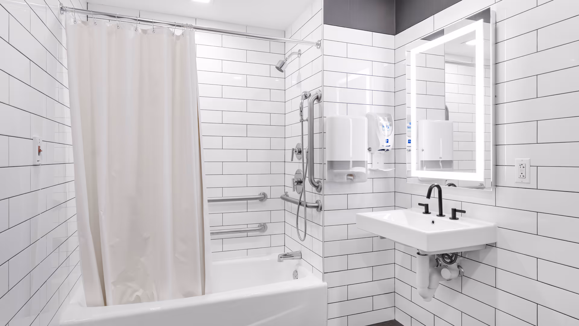 Accessible white-tiled bathroom with bathtub/shower, multiple grab bars, a wall-mounted sink, and an illuminated mirror.