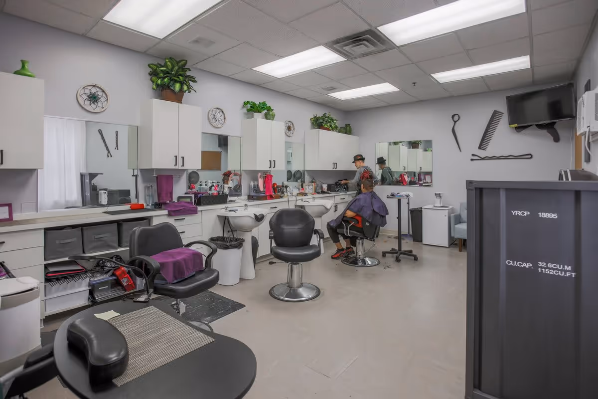 Interior of a hair salon with white walls and ceiling lights. There are multiple black salon chairs, sinks, and white cabinets with plants on top. A person is seated in one of the salon chairs getting a haircut from a stylist. Hairdressing tools and products are visible on the counters. Wall decorations include large scissors, a comb, and hairpins.