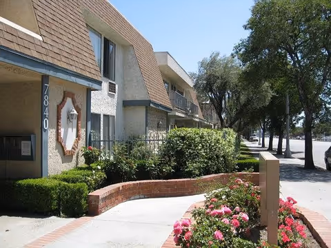 Exterior view of a senior apartment building with beige walls and brown shingled roof accents. The building number 7840 is visible near the entrance. A curved brick planter filled with green shrubs and pink flowers lines the sidewalk leading to the entrance. Trees and a street are visible in the background under a clear blue sky.