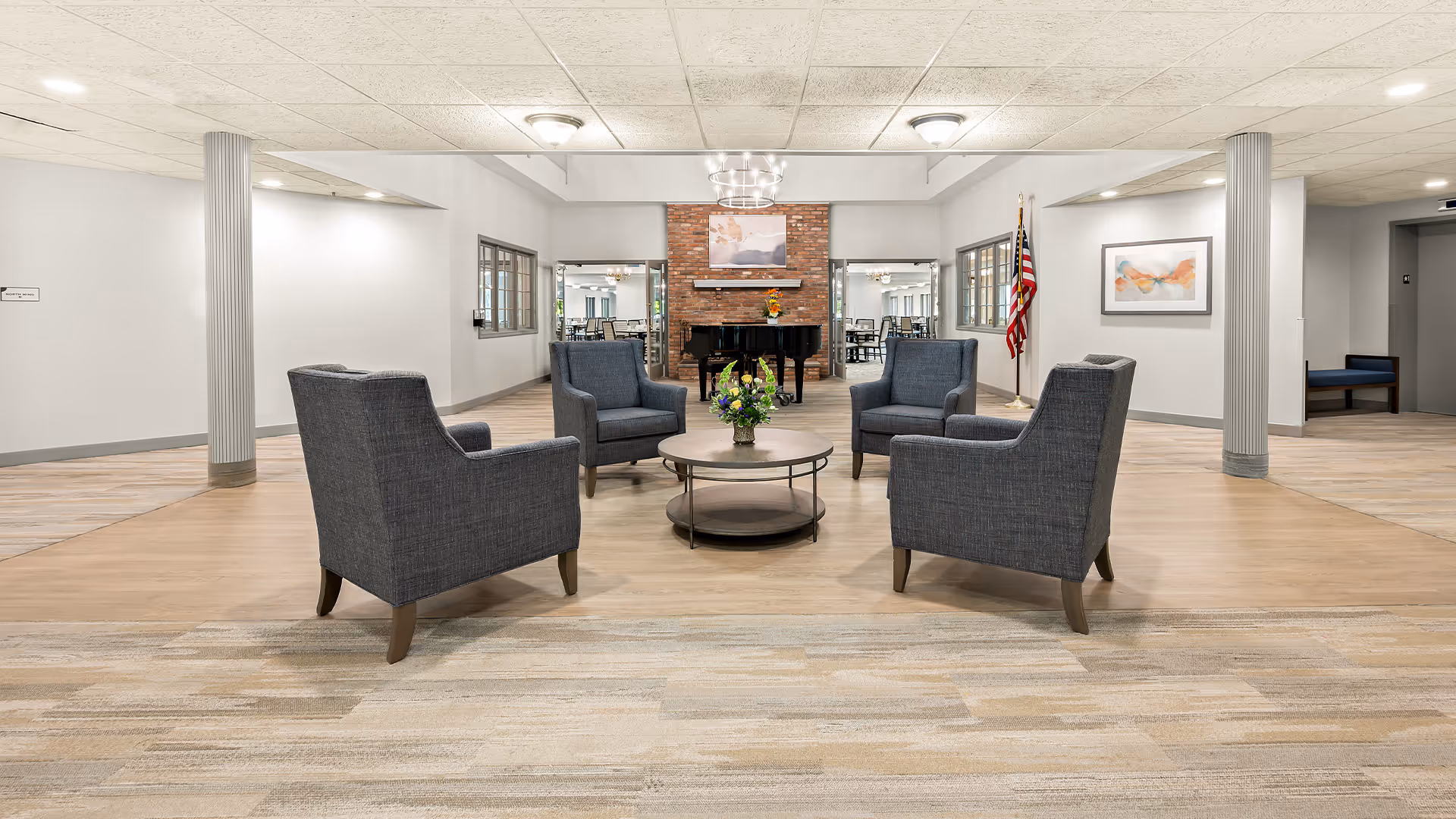 A spacious and well-lit common area with four gray upholstered armchairs arranged around a round coffee table with a flower arrangement. In the background, there is a brick wall with a black grand piano and a framed artwork above it. The room has light-colored walls, carpeted and wooden flooring, two pillars, and an American flag to the right side.
