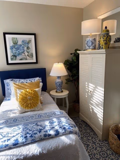 A cozy bedroom with a single bed featuring blue and white floral bedding, a yellow decorative pillow, and a white pillow. Next to the bed is a small round white side table with a blue and white patterned lamp. A large white wardrobe stands beside the table, with sunlight casting shadows on it. A framed floral artwork hangs on the beige wall above the bed, and a green potted plant is placed between the wardrobe and the wall.
