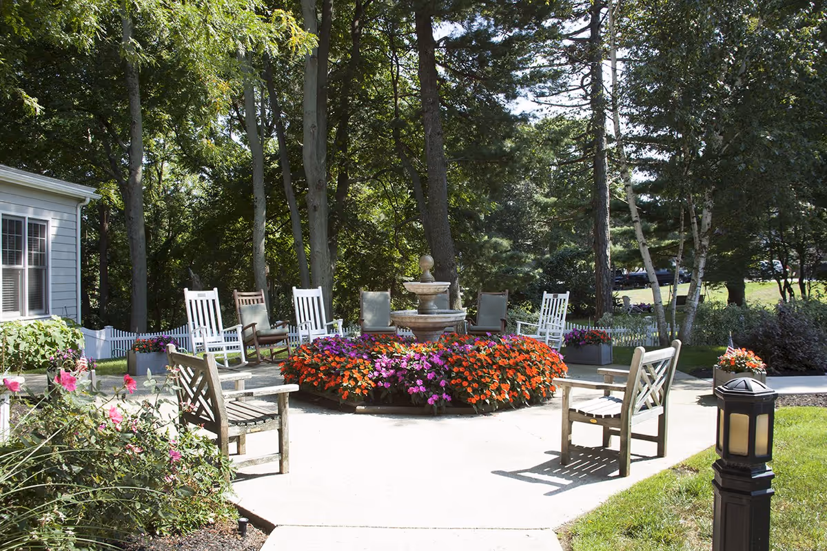 Outdoor seating area in a garden with wooden benches and white rocking chairs arranged around a circular flower bed with colorful flowers and a stone fountain in the center, surrounded by trees and greenery.