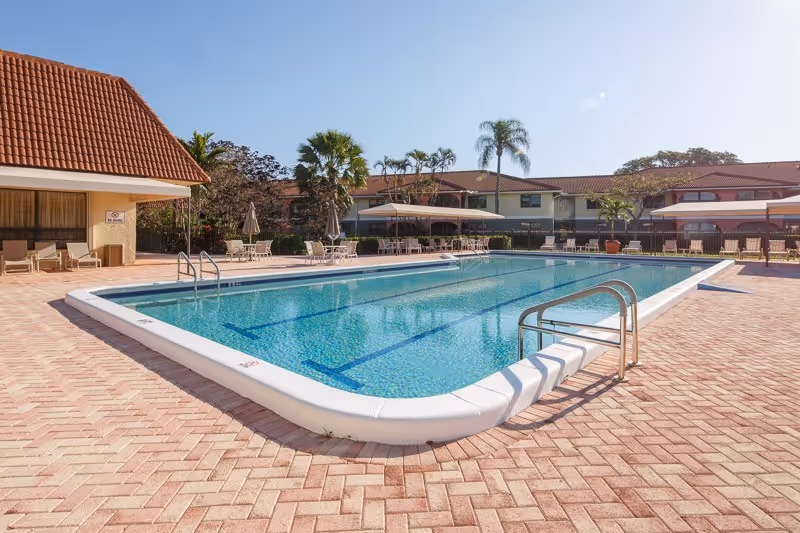 A clean outdoor swimming pool with lounge chairs, umbrellas, palm trees and two-story facility buildings under a clear sky.