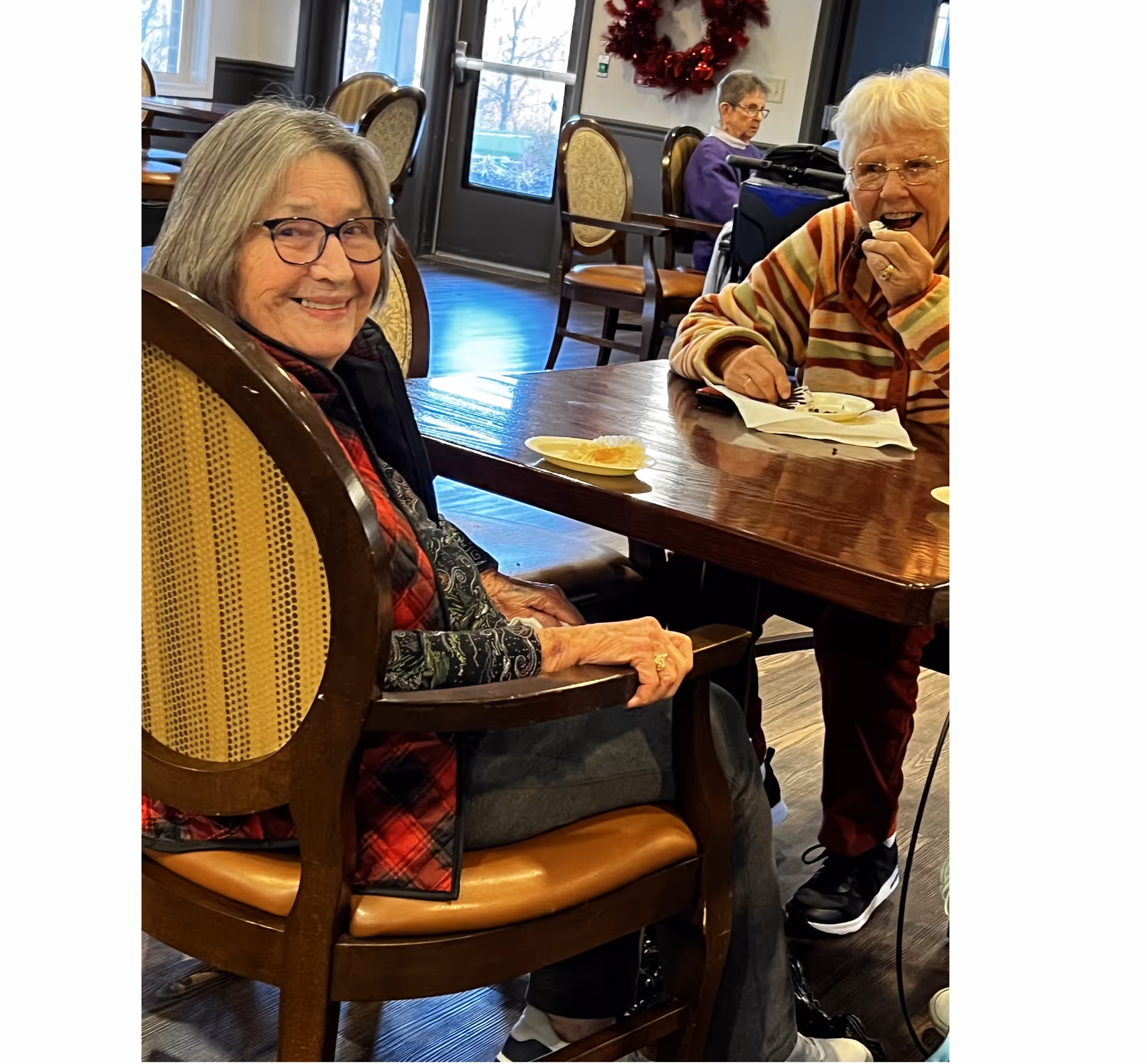 Two elderly women sitting at a wooden table in a communal dining area, one smiling at the camera and the other eating a dessert. In the background, another elderly woman is seated near a door with a festive wreath on the wall.