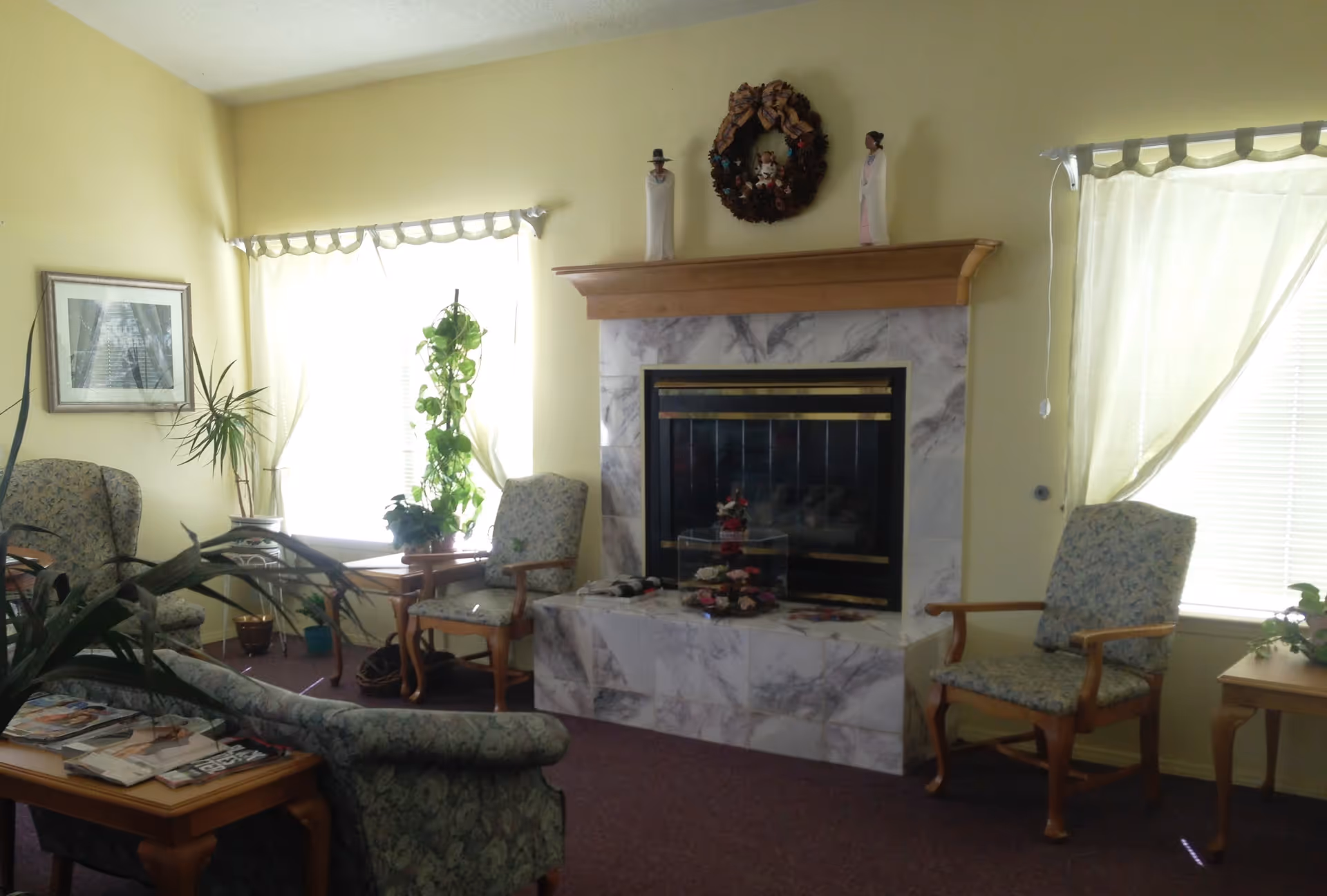 A cozy living room with a marble fireplace decorated with a wreath and figurines on the mantle. The room has yellow walls, two large windows with light curtains, several upholstered chairs with wooden arms, a wooden coffee table with magazines, and various potted plants.