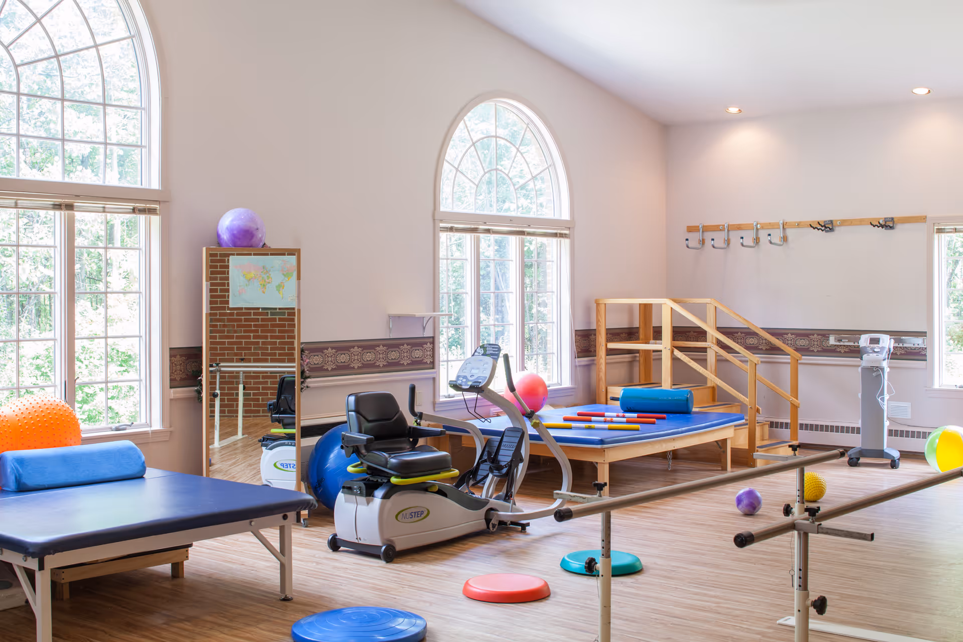 Bright physical therapy room with large arched windows letting in natural light. The room contains exercise equipment including a recumbent bike, parallel bars, therapy tables with colorful exercise balls and foam rollers, and a wooden staircase for rehabilitation exercises. The walls are light-colored with decorative trim and hooks for hanging items.