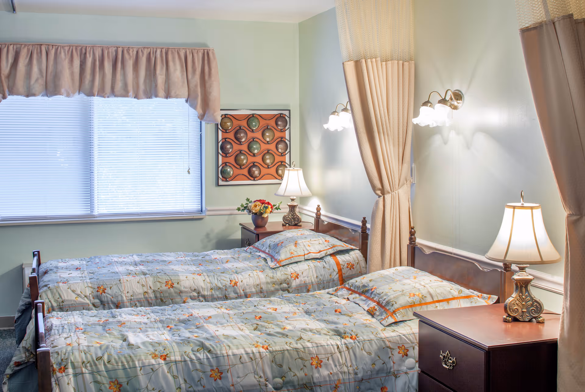 A bedroom with two twin beds side by side, each with floral patterned bedding. There are two bedside tables, each with a decorative lamp. A window with blinds and a valance is on the left wall, and a framed decorative piece hangs between the beds. Light-colored curtains hang on the right side of the beds.