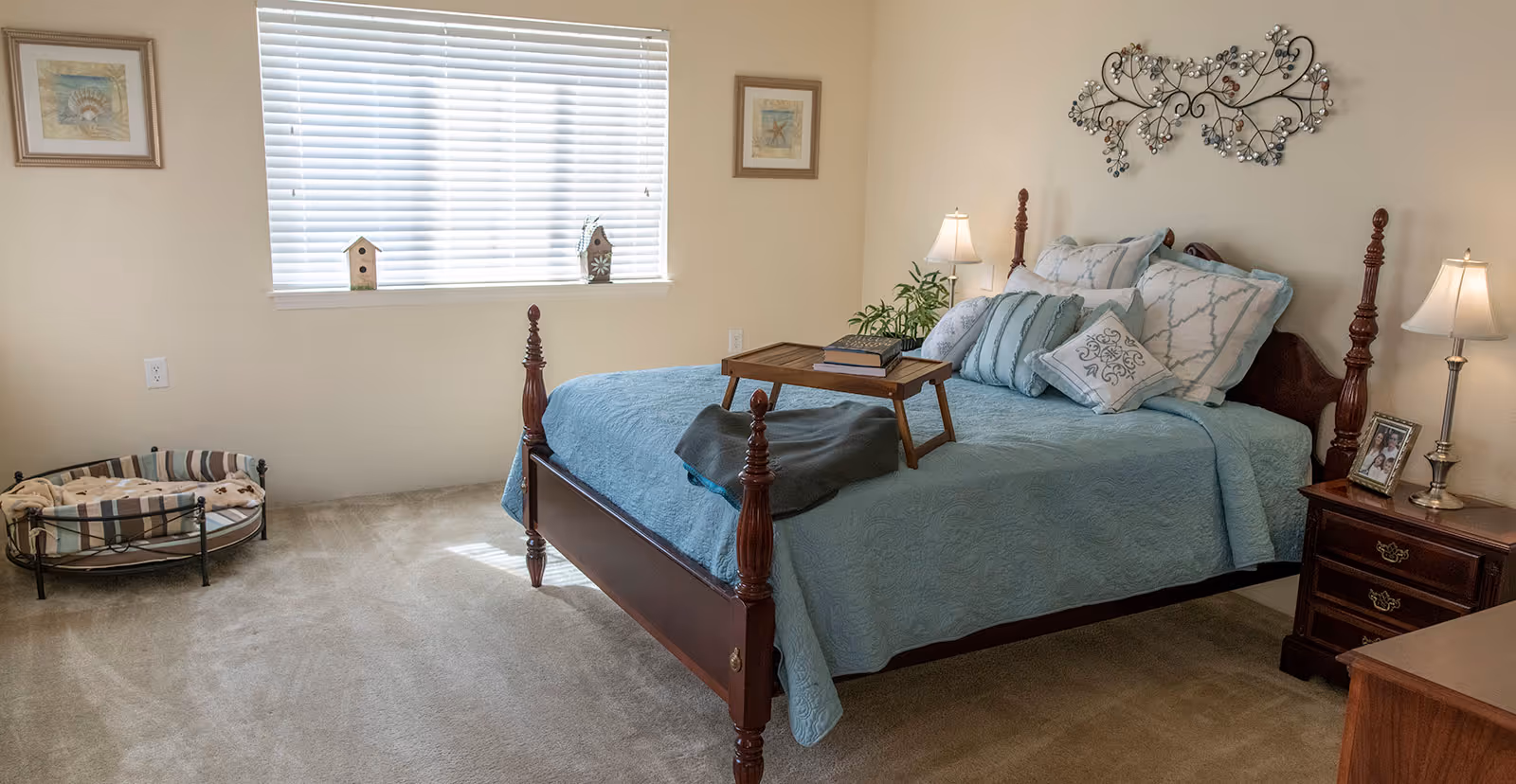 A cozy bedroom with a wooden four-poster bed covered in a light blue quilt and multiple pillows. A wooden tray with books and a folded blanket rests on the bed. There are two bedside tables with lamps and a framed photo on the right side. The room has beige walls with two framed pictures and a decorative metal wall art above the bed. A window with white blinds lets in natural light, and a pet bed is placed on the carpeted floor near the window.