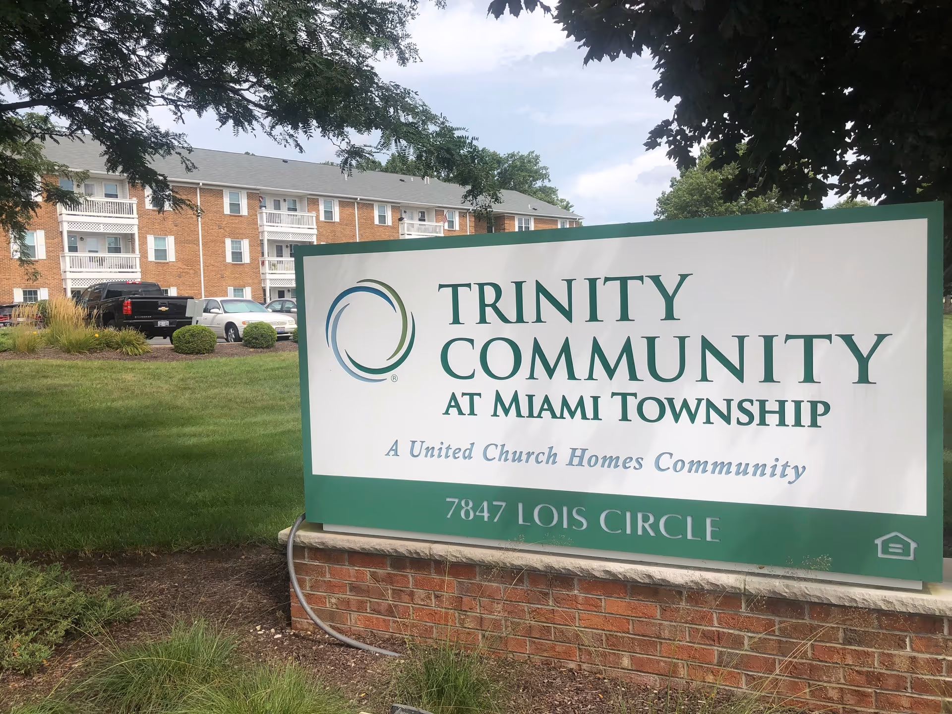 Outdoor view of a senior living facility sign that reads 'Trinity Community at Miami Township, A United Church Homes Community, 7847 Lois Circle' with a brick building and parked cars in the background under a partly cloudy sky.