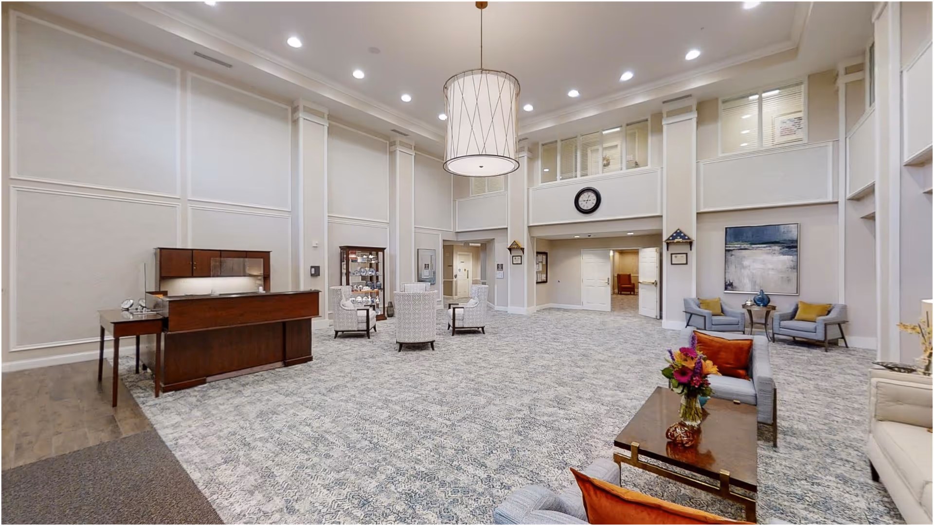 Spacious and well-lit common area with high ceilings, featuring a large hanging light fixture, multiple seating arrangements with chairs and sofas, a wooden reception desk, a display cabinet, a wall clock, and decorative artwork on the walls.