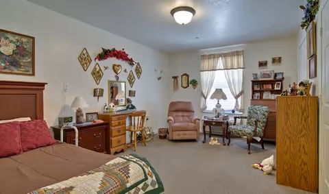 A cozy senior living bedroom featuring a bed with a quilted blanket and burgundy pillows, a wooden dresser with a mirror decorated with floral and heart-shaped wall hangings, a desk with a chair, a recliner, two armchairs near a window with sheer curtains, and a wooden cabinet. The room is warmly lit with a ceiling light and a table lamp.
