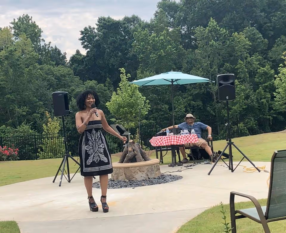 A woman in a black and white strapless dress is speaking or singing into a microphone outdoors on a concrete patio. Behind her, a man wearing a hat and sunglasses is seated at a table covered with a red and white checkered tablecloth under a blue umbrella. There are two large speakers on stands on either side of the patio, and green trees and grass surround the area.