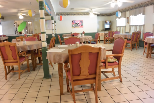 A dining room with multiple round tables covered with beige tablecloths and surrounded by wooden chairs with red cushions. The room has tiled flooring, ceiling fans, and windows with valances. Colorful hanging decorations are visible, and a beverage dispenser is in the back corner.