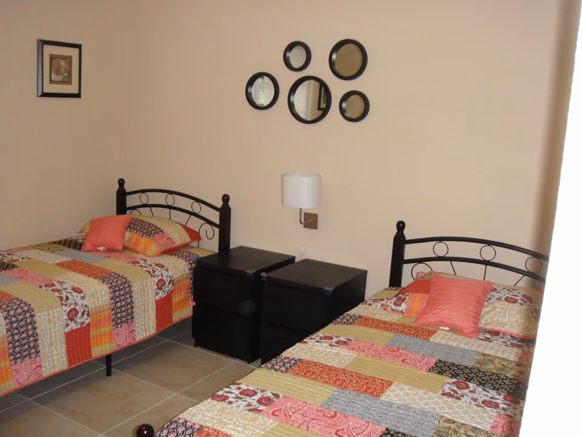 A bedroom with two twin beds featuring black metal headboards and colorful patchwork quilts in shades of red, orange, yellow, and black. Each bed has a pink pillow. Between the beds are two black nightstands with a white wall-mounted lamp above them. On the wall above the lamp are five round mirrors arranged in a decorative pattern. A small framed picture hangs on the adjacent wall.